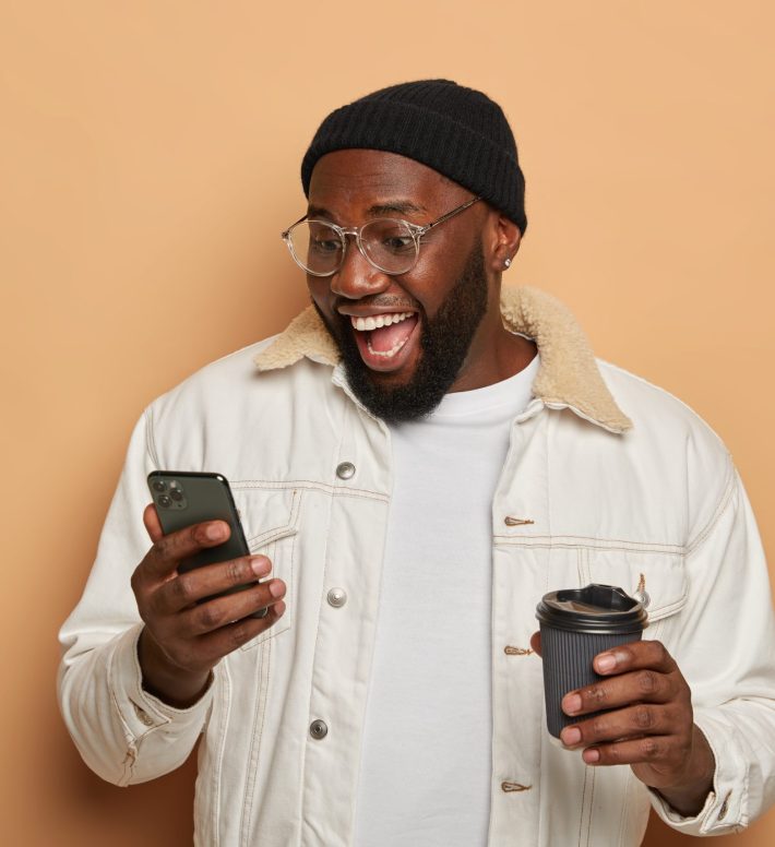 Happy man with thick beard, dark skin, enjoys online chatting via modern cellular, gets pleasant unexpected message or funny sms, drinks takeaway coffee, wears stylish clothes, poses indoor. Happy man with thick beard, dark skin, enjoys online chatting via modern cellular, gets pleasant unexpected message or funny sms, drinks takeaway coffee, wears stylish clothes, poses indoor.