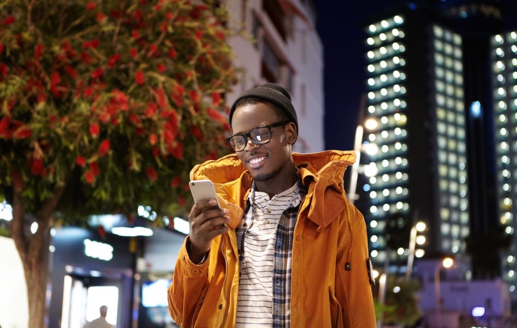 Handsome young dark-skinned hipster in hat, eyewear and winter coat messaging his girlfriend online on smart phone while waiting for her on street at night, looking at screen with joyful smile
