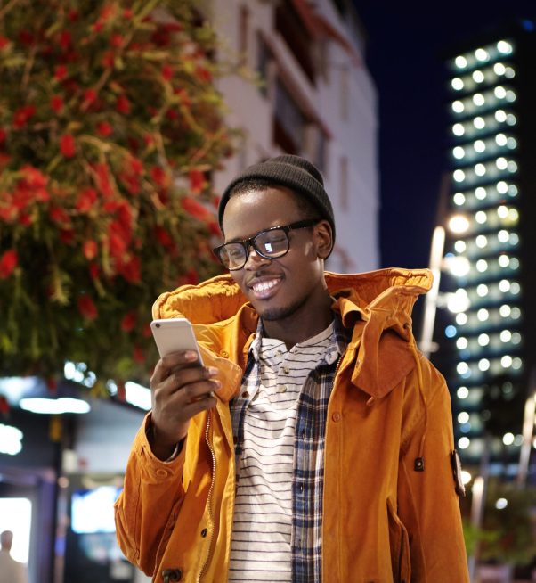 Handsome young dark-skinned hipster in hat, eyewear and winter coat messaging his girlfriend online on smart phone while waiting for her on street at night, looking at screen with joyful smile